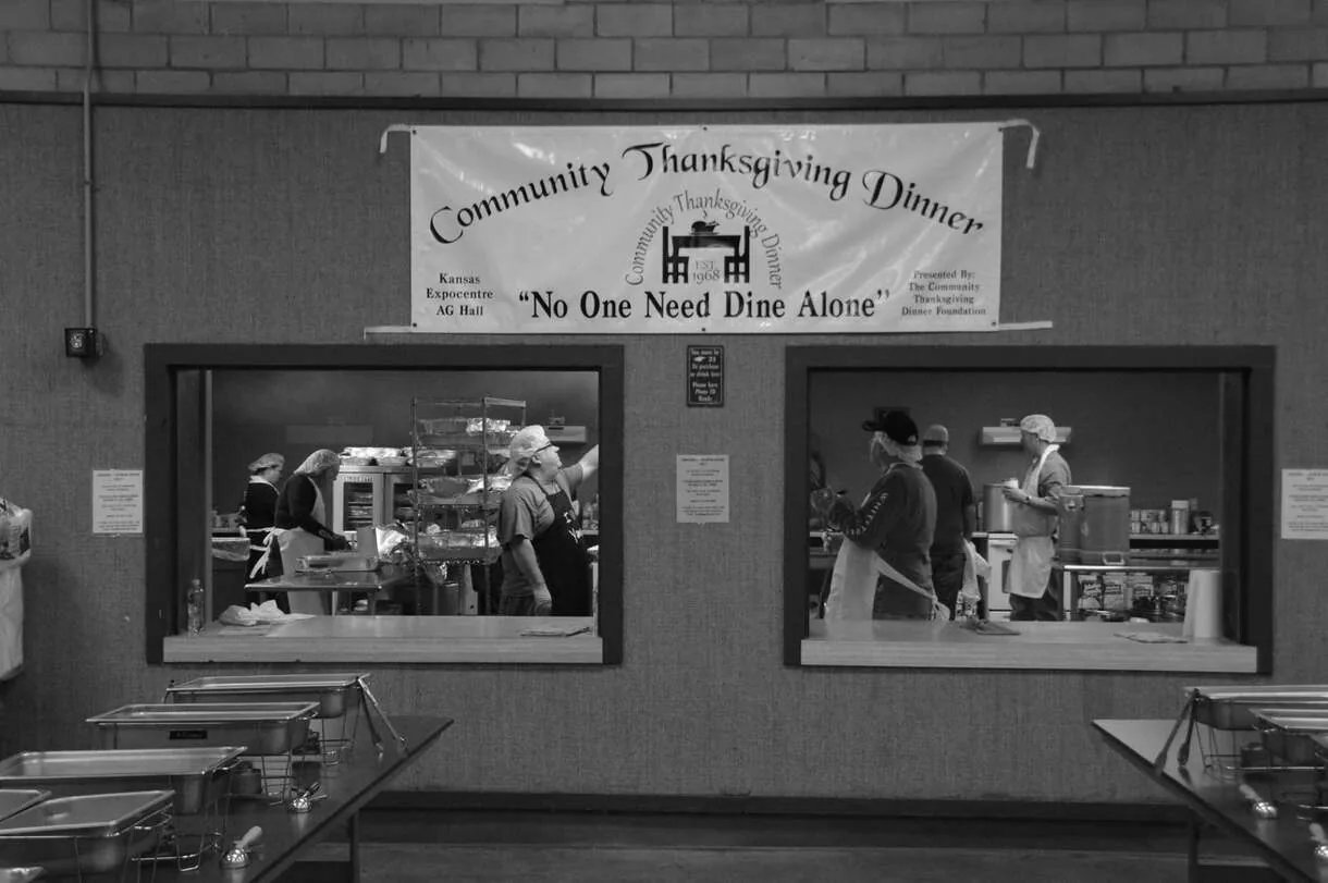 Black-and-white photograph of volunteers behind two serving windows beneath a Community Thanksgiving Dinner banner reading No One Need Dine Alone.